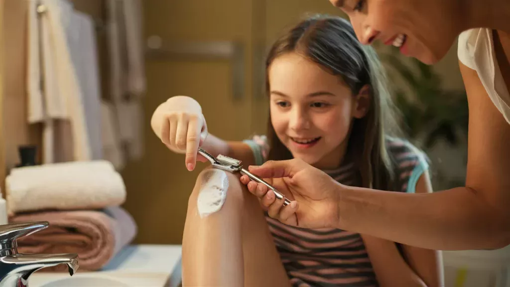 Mom teaching her daughter how to shave her legs for the first time with a manual razor in the bathroom.