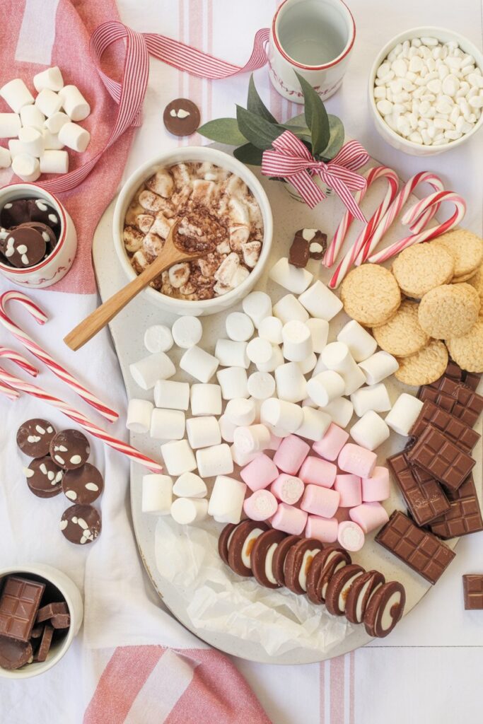 Hot cocoa and sweets board filled with marshmallows, chocolate bars, cookies, peppermint sticks, and a bowl of cocoa topped with marshmallows, styled with festive ribbons and a red-striped cloth for a cozy Christmas setup.