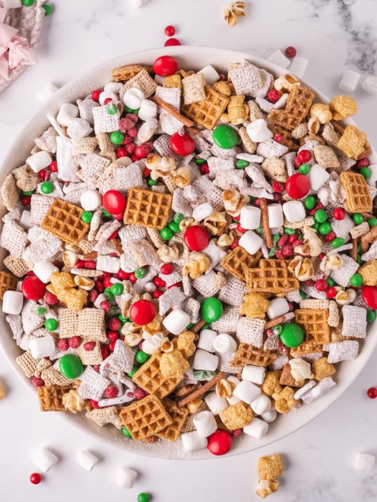 A bowl filled with colorful Christmas snack mix made of Chex cereal, mini marshmallows, pretzels, waffle cereal, popcorn, and red and green candies, dusted with powdered sugar on a white marble surface.