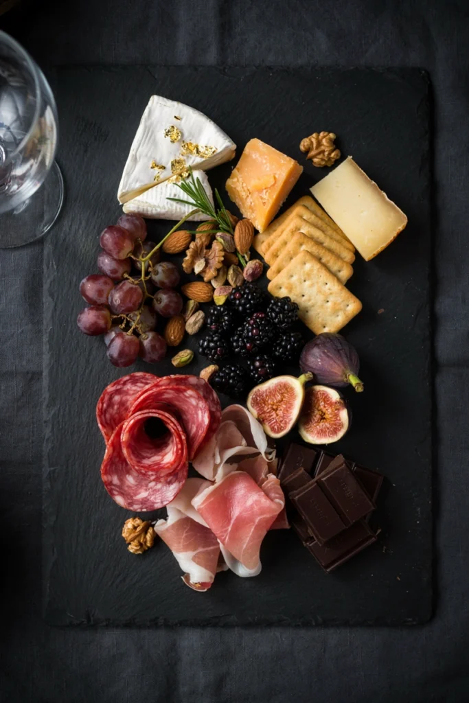 A black slate board styled with dark cheeses, black grapes, gold-rimmed crackers, and edible gold leaf over a wedge of brie
