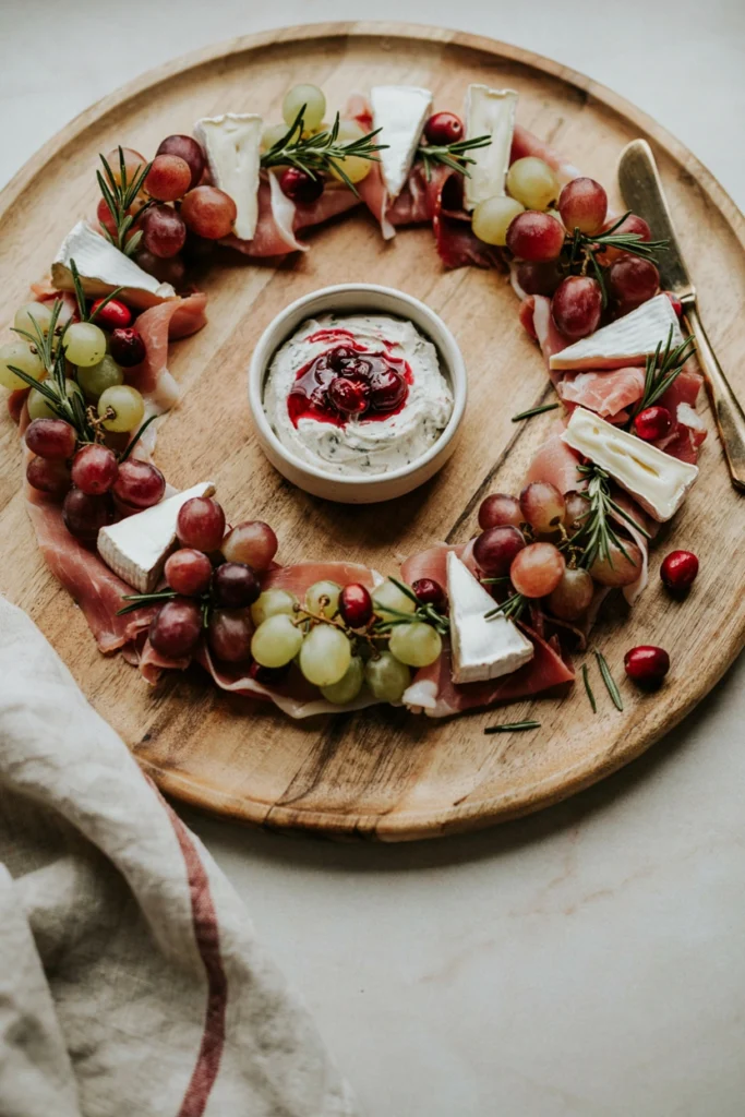 A circular charcuterie board arranged as a wreath with a bowl of cranberry sauce in the center, ringed by prosciutto ribbons and fresh herbs