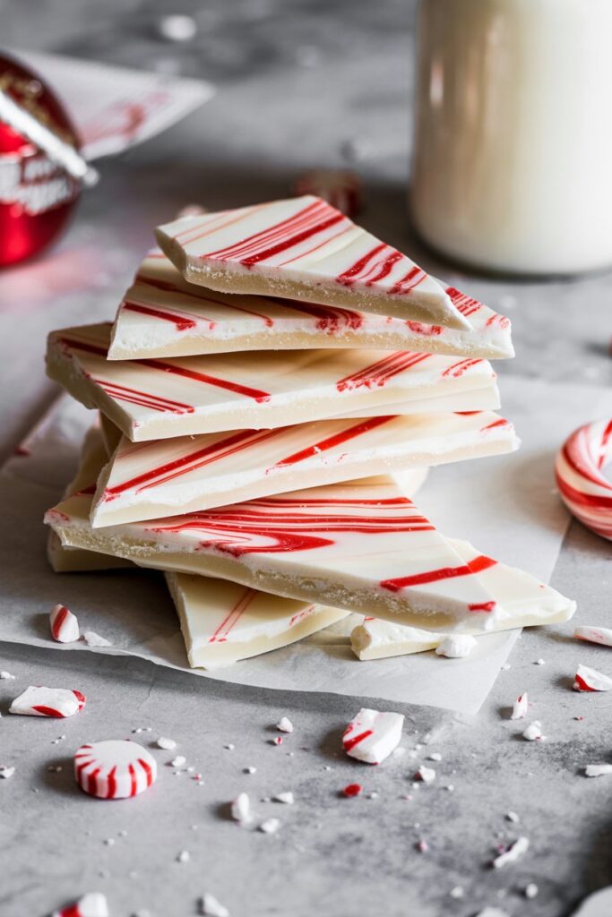 Stack of white chocolate peppermint bark with red candy cane swirls on parchment paper, surrounded by crushed peppermint pieces and festive holiday decor in soft lighting.