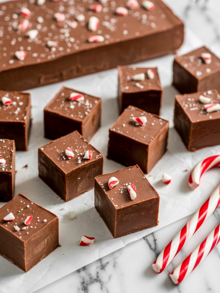 Squares of rich chocolate fudge topped with crushed candy canes and sea salt, arranged on parchment paper with candy canes beside them on a marble surface for a festive Christmas treat.