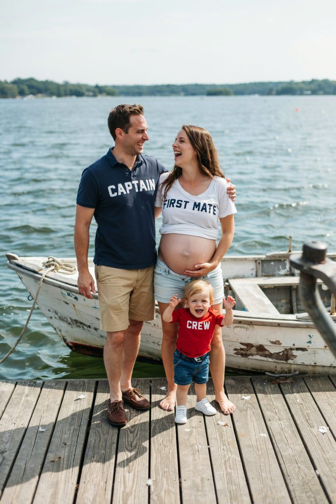 A family — dad in a "Captain" polo, mom in a "First Mate" shirt with her bump visible, and toddler in a "Crew" tee — standing at the bow of a small boat or dock with water behind them. Everyone is grinning. Casual, bright midday light. No formal styling.