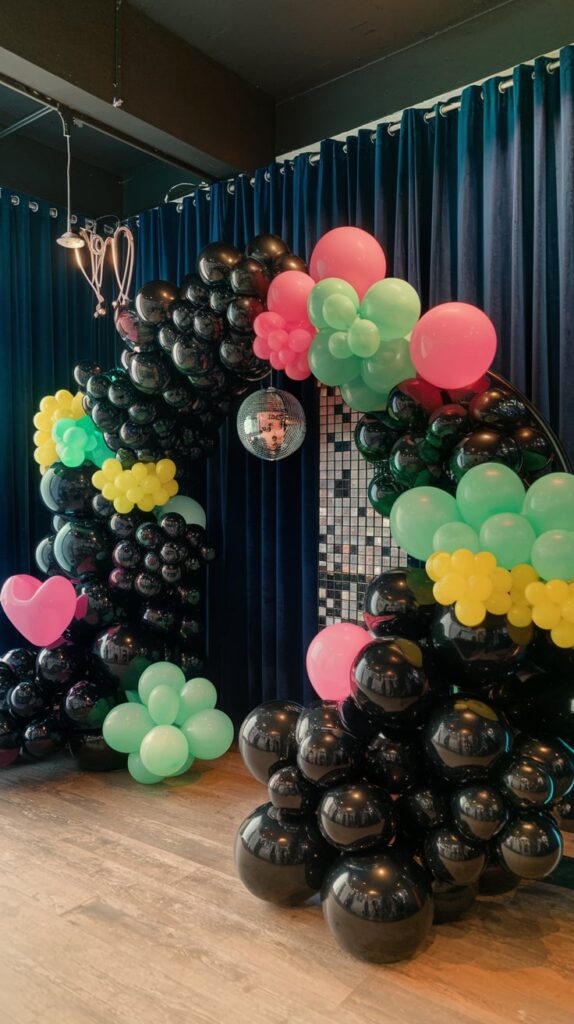 Black balloon garland arch accented with pink, green, and yellow balloon clusters and a disco ball