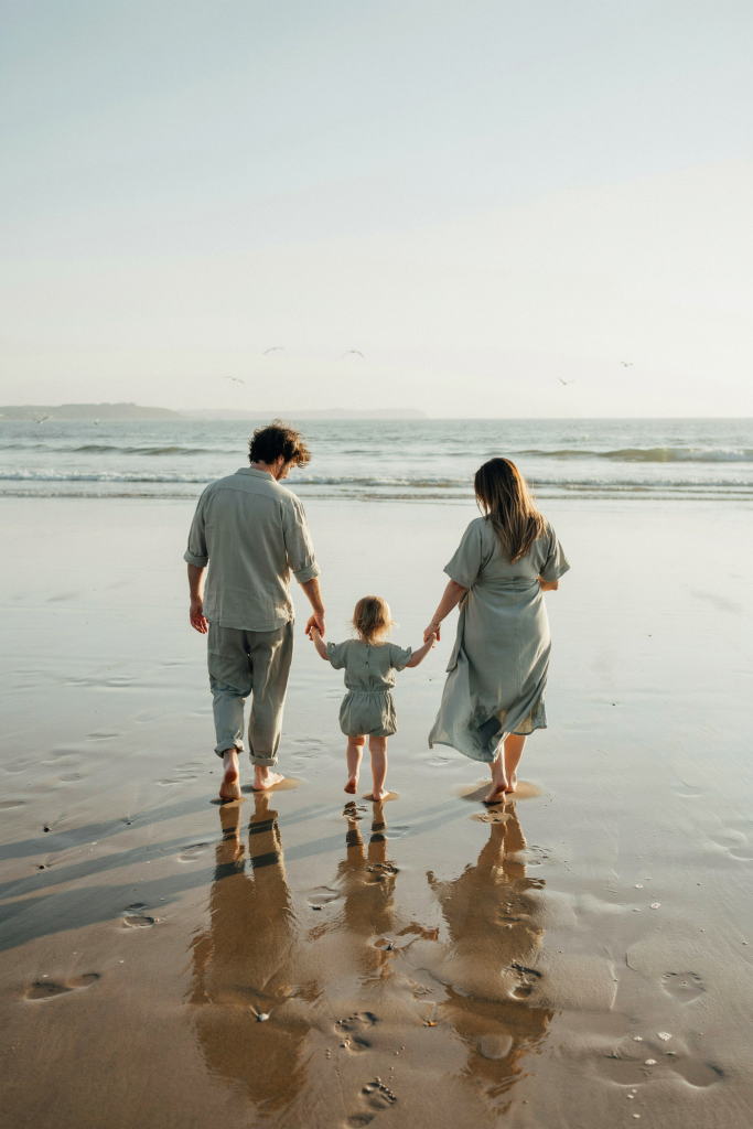 A family of three in matching sage-green linen — dad, toddler daughter, and visibly pregnant mom — walking barefoot on the beach away from the camera, holding hands. Aerial or slightly elevated angle. Warm afternoon light. No text overlay, no props