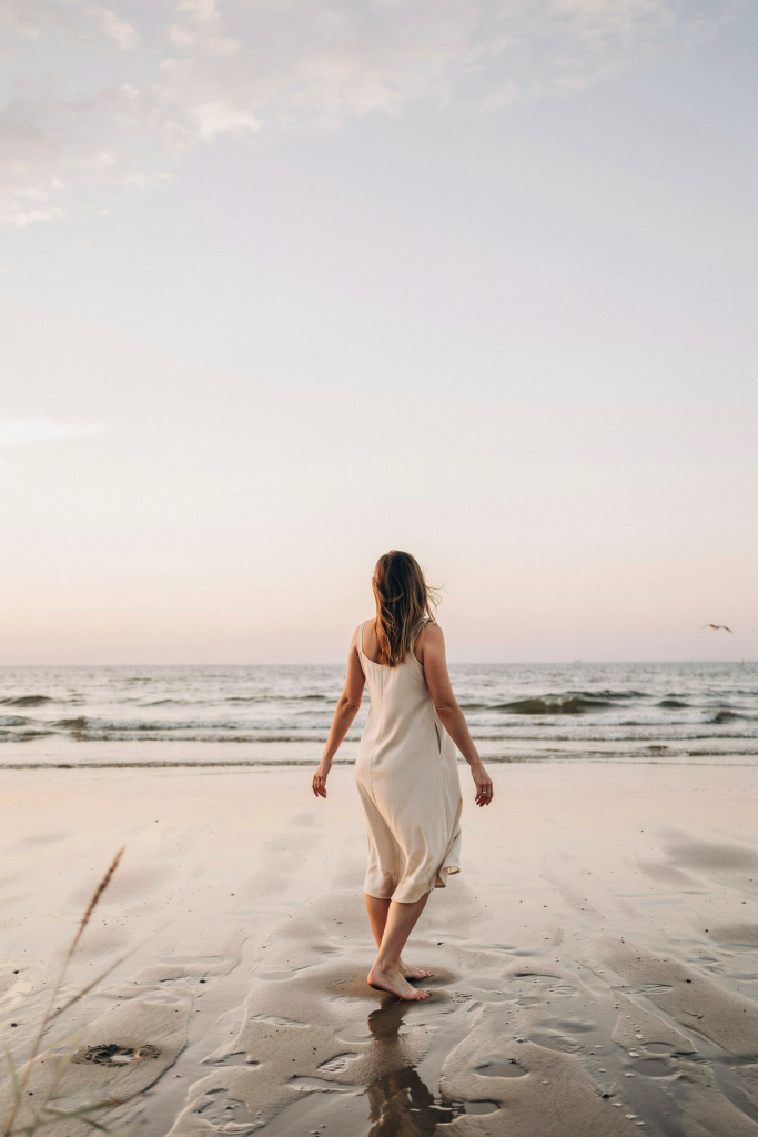 A woman standing alone at the water's edge at sunset, facing away from the camera. She's visibly pregnant, barefoot, arms relaxed at her sides, looking out to sea. The sky is a pale gold and lavender gradient. No text. No props. No other people. Just her, the water, and whatever she's thinking about right now.