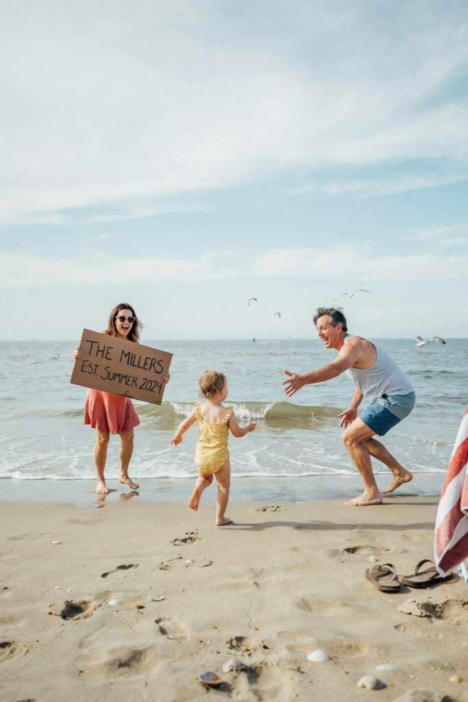 An informal, slightly blurry beach photo where the toddler has clearly abandoned the planned pose and is running toward the water, parents laughing in the background — one reaching out to grab the child, the other holding a sign that has gone slightly askew. Everything is chaos. Everything is perfect. Shot candidly with no particular composition intent.