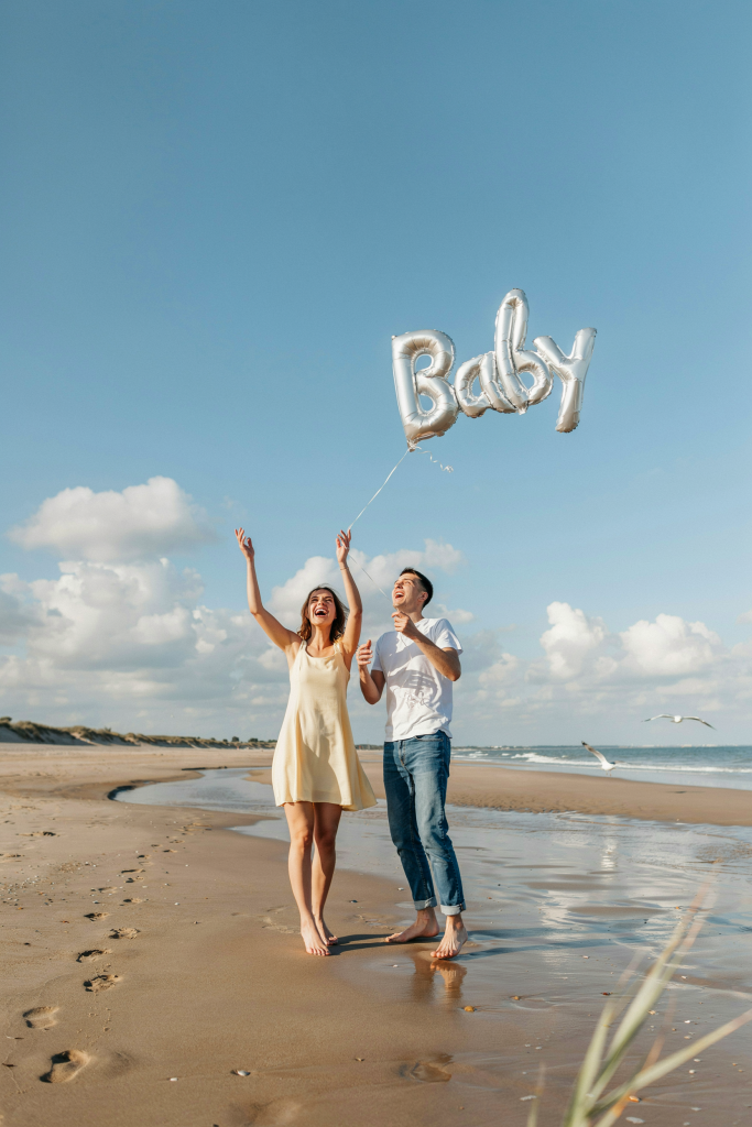 A couple on the beach releasing a single oversized "Baby" foil balloon into the sky, hands raised and laughing, the balloon trailing upward against a blue cloud-dotted sky. Both dressed casually — jeans and a sundress. Mid-afternoon light. Completely spontaneous-looking.