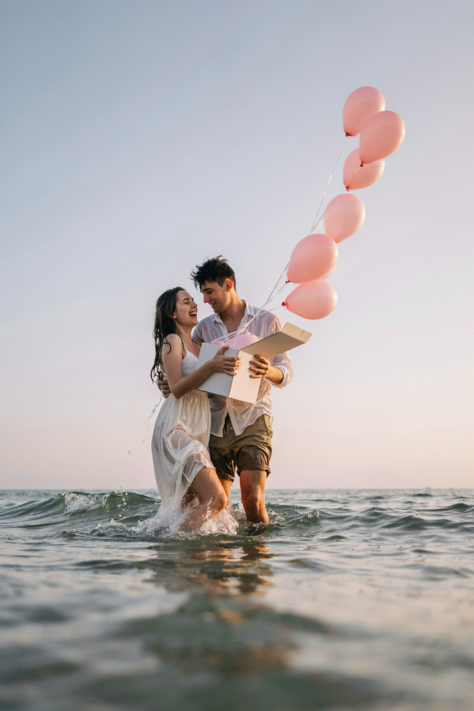 A young couple stands knee-deep in the ocean, holding an open white box above the waves. Pink balloons escape upward into the blue sky. The look on her face — pure joy, mouth open, leaning into him — is the image. Shot from the side at a slight distance. Pastel sky, warm late afternoon light.