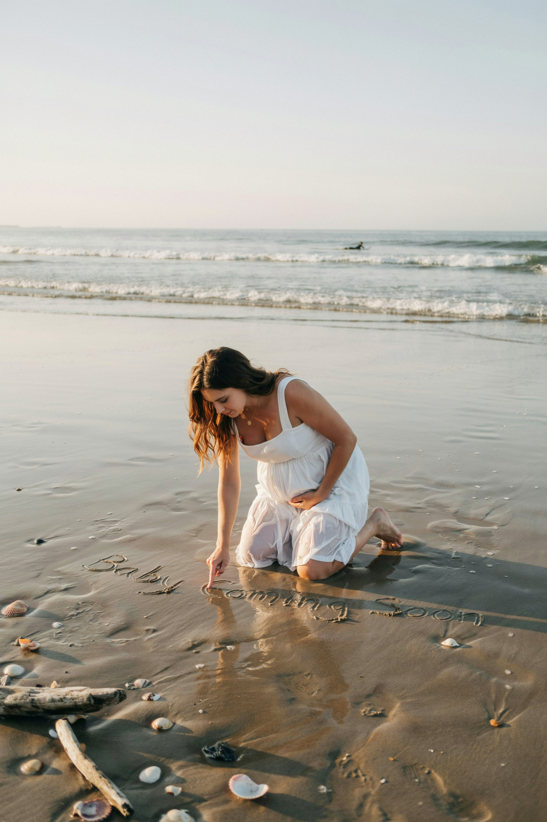 miling pregnant woman in a flowy white sundress kneeling in wet sand, writing "Baby Coming Soon" in the sand as waves lap behind her.