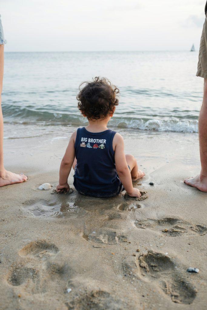 A toddler in a navy "Big Brother" tank top sitting in the sand at the water's edge, looking out to sea. Shot from behind — curly hair, chunky toddler legs, small fists in the sand. Parents' legs just visible on either side. Candid, completely unposed.