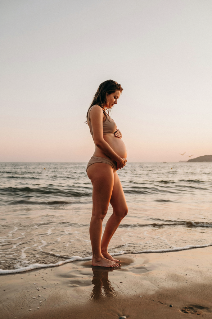 A side-profile shot of a pregnant woman in a bikini standing ankle-deep in gentle surf, both hands cradling her belly. Shot at golden hour — warm orange and pink tones on the water. Completely natural, no props, no text overlay.