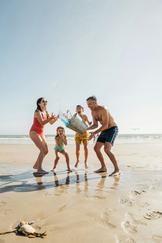 A family of four — mom, dad, and two kids — standing in a circle on the beach around a large bucket. They tip the bucket and a cascade of bright blue water splashes outward, catching the light. All four are screaming and laughing. Mid-motion shot — frozen water droplets everywhere. Bright afternoon light.