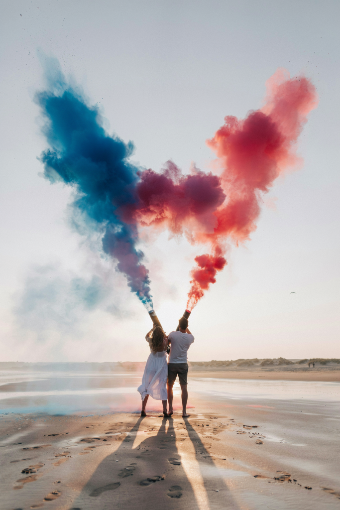 A couple on an empty sunrise beach firing two large powder cannons — one releasing blue powder, the other pink — simultaneously, the clouds of color meeting in the middle above their heads. Shot from behind. Sun barely visible on the horizon. Long, dramatic shadows in the wet sand in front of them