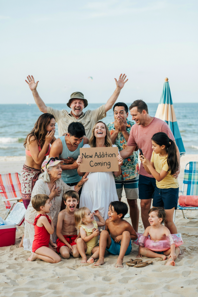 The whole extended family crammed into a beach photo — grandparents, aunts, uncles, cousins, everyone. Mom and dad are in the center; mom is clearly pregnant and holding a sign that reads "New Addition Coming." Everyone is laughing, multiple people are covering their mouths in shock, one grandma is crying. Completely spontaneous. Beach chairs and towels everywhere in the background.