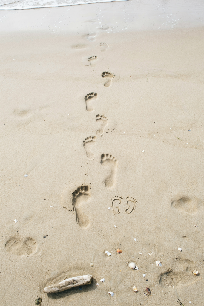 Three sets of adult footprints — two large, one child-sized — walking toward the ocean in damp sand, with one tiny set of bare baby feet drawn in the sand beside the smallest set. Shot from above. The ocean is just visible at the top of the frame.