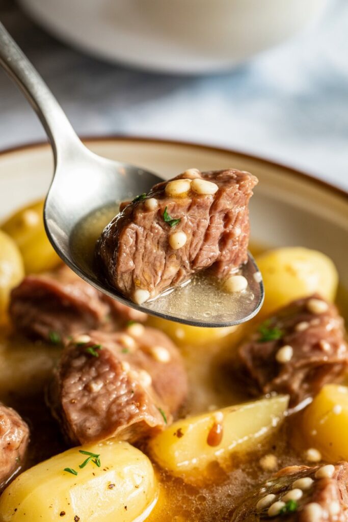A close-up of a spoon lifting a tender garlic butter beef bite coated in glossy garlic butter sauce, with baby potatoes visible beneath and a softly blurred background.