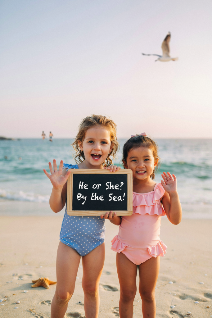 Two children — one in a blue polka-dot swimsuit, one in a pink ruffle two-piece — hold a chalkboard sign between them on the beach that reads "He or She? By the Sea!" Waving at the camera. Both have their gender color repeated in their swimsuits. Golden hour light, slightly backlit.
