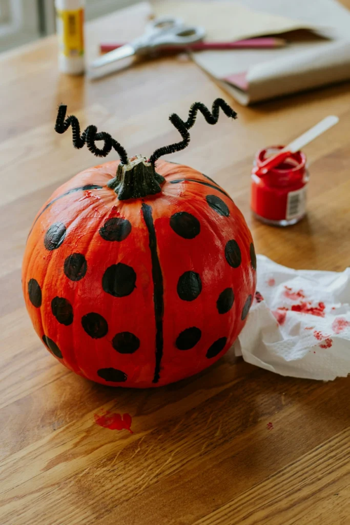 Red pumpkin painted as a ladybug with black dots and pipe cleaner antennae
