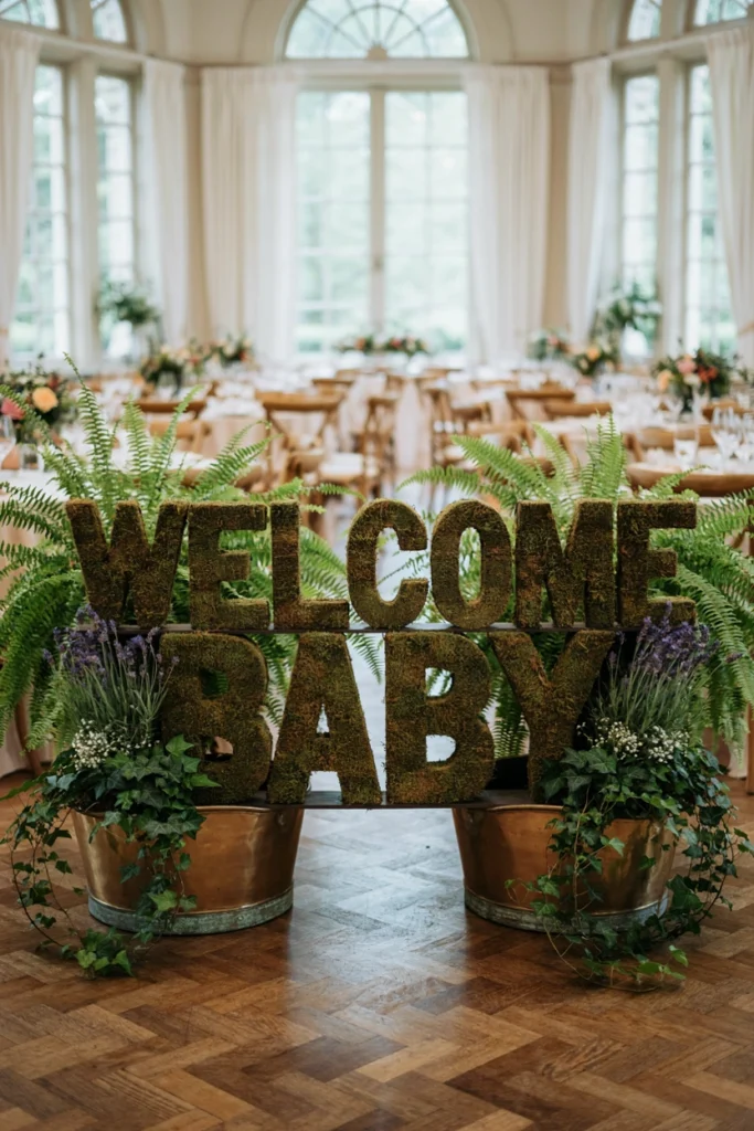 Large moss-covered letters spelling Welcome Baby propped against greenery