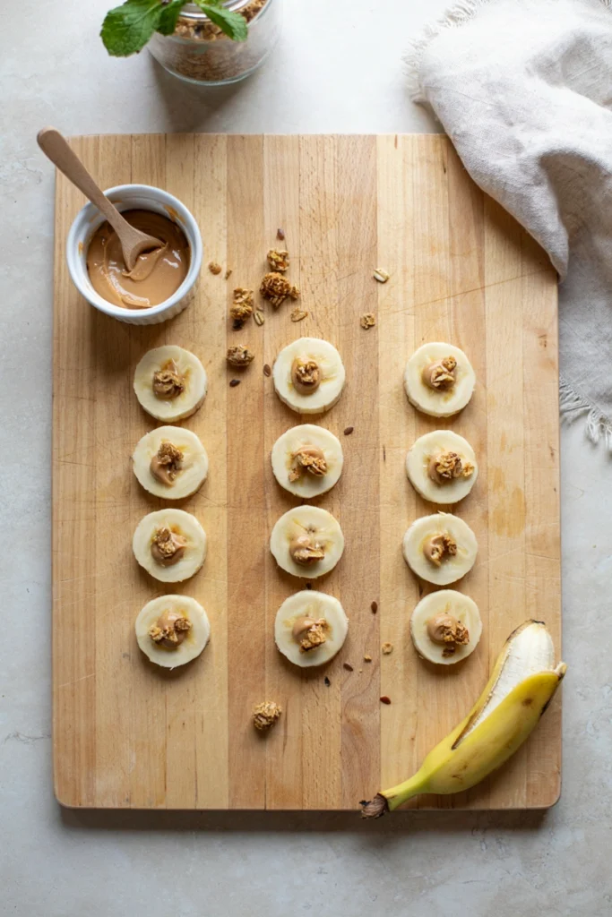Banana rounds topped with a swirl of peanut butter and a mini granola sprinkle, arranged in neat rows on a wooden board