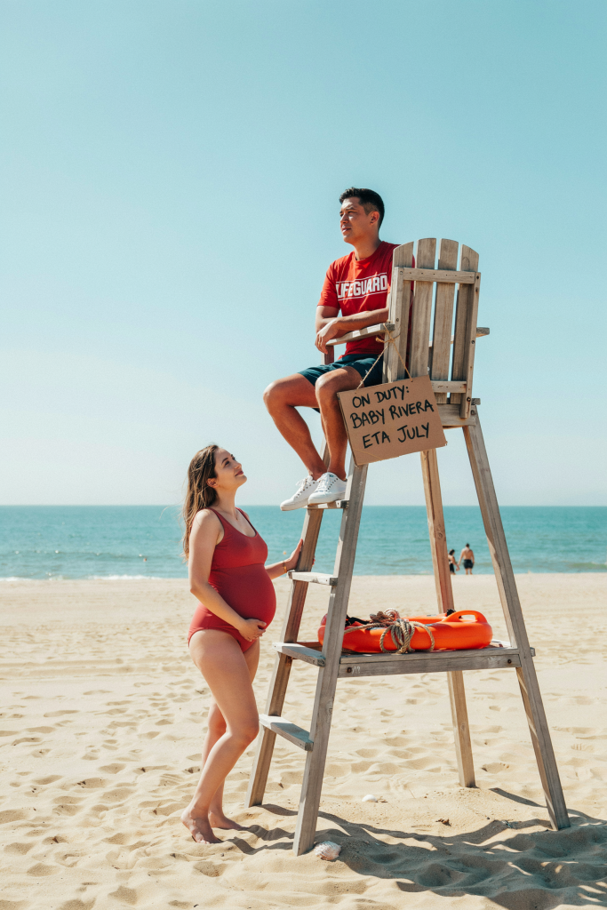 Dad in a red "Lifeguard" t-shirt sits in an actual lifeguard chair, arms folded, looking out to sea. Mom stands at the base of the chair, hand on the ladder, visibly pregnant in a red swimsuit. A small handmade sign hangs from the chair reading "On Duty: Baby [Last Name] — ETA [Month]." Bright midday light, very graphic and bold composition.
