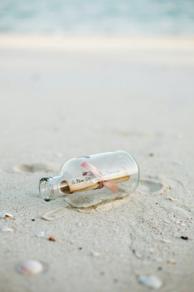 A clear glass bottle half-buried in white sand with a rolled scroll of paper inside tied with a blush ribbon. The scroll reads "A New Arrival is Coming." Shot at a low angle with water and horizon blurred in the background. Warm, slightly overcast light.