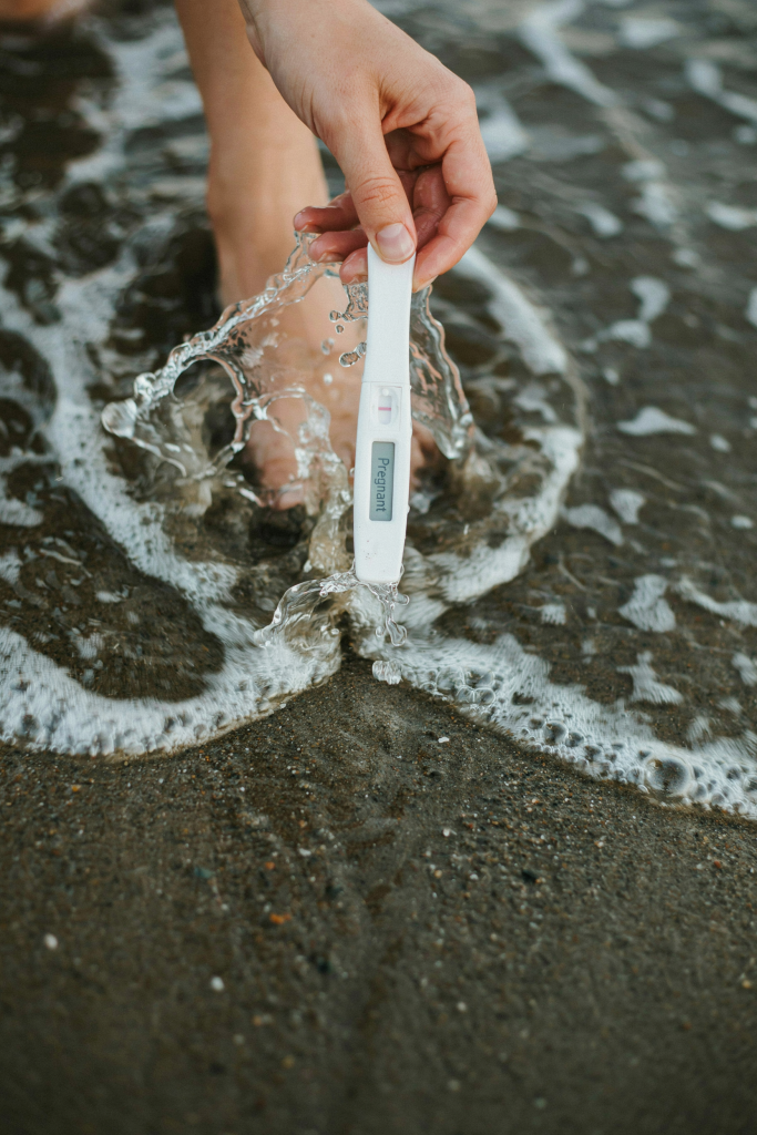 A woman's hand holding a positive pregnancy test just at the water's edge, waves lapping around her wrist. Shot from above — just the hand, the test, and the foamy white wave. No face, no body, just the test and the ocean. Clean, minimal, instantly understood.