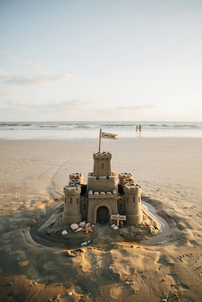A detailed sand castle about 18 inches tall with multiple towers. A small flag at the tallest tower reads "Coming Soon" and a tiny sign at the gate reads "Baby [Last Name]." Built on dry sand with a moat. Shot at a low angle so the castle is in the foreground and the ocean stretches to the horizon behind it. Afternoon light, shadows giving the castle depth