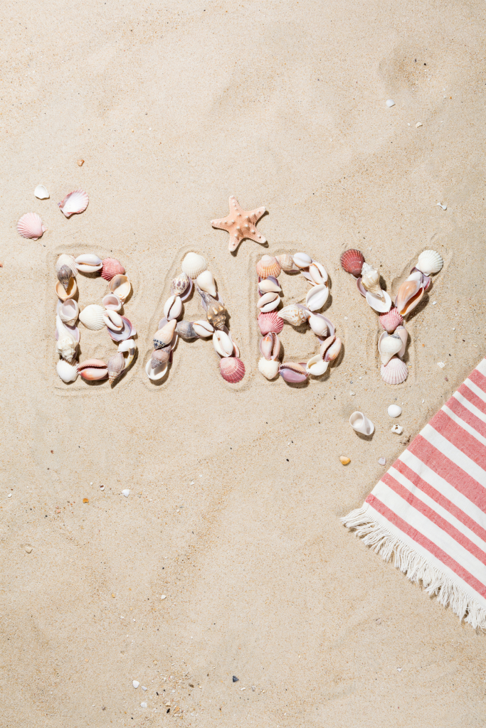A flat-lay close-up of seashells arranged on dry sand to spell "BABY" in thick block letters. Mix of pink, white, and peach shells. Shot from directly above with a slight overhead shadow giving depth. Beach towel edge visible in lower right corner.