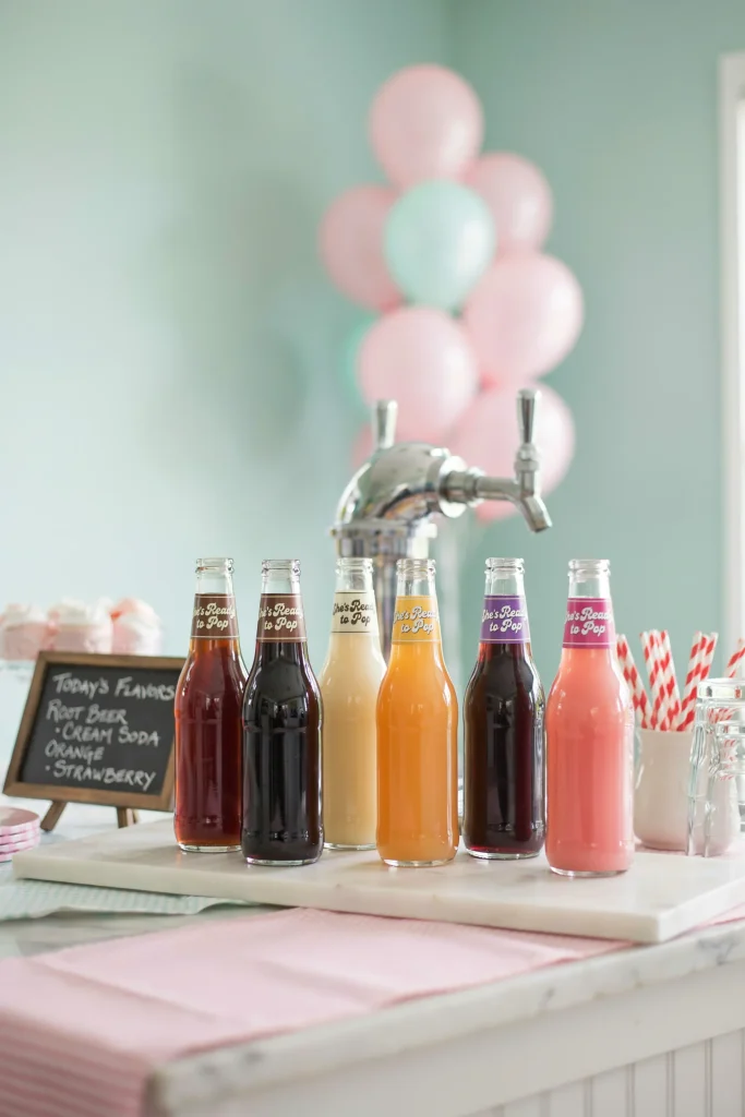 A beverage station displaying glass bottles of colorful sodas (root beer, cream soda, orange soda, grape soda, strawberry) with custom "She's Ready to Pop" labels wrapped around each bottle neck. Fun, retro, playful vibe.