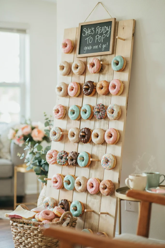 A wooden or metal donut wall holding 20-30 glazed, frosted, and sprinkle-covered donuts in coordinating colors. A sign reads "She's Ready to Pop" or a donut-themed pun. Guests can grab donuts throughout the party.