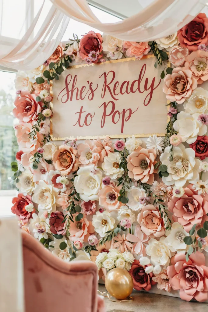 A flower-covered backdrop featuring oversized paper flowers in soft pastels and blush tones, with a large "She's Ready to Pop" or "Welcome Baby" sign positioned prominently. Lighting is soft and flattering. Photo-ready moment