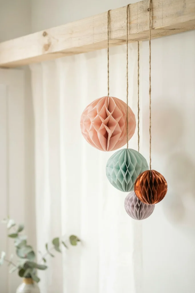 Three or four colorful honeycomb balls (tissue paper spheres) hanging at different heights from a ceiling beam or decorative string. Pastel colors mixed with metallics. Simple, elegant, and whimsical.