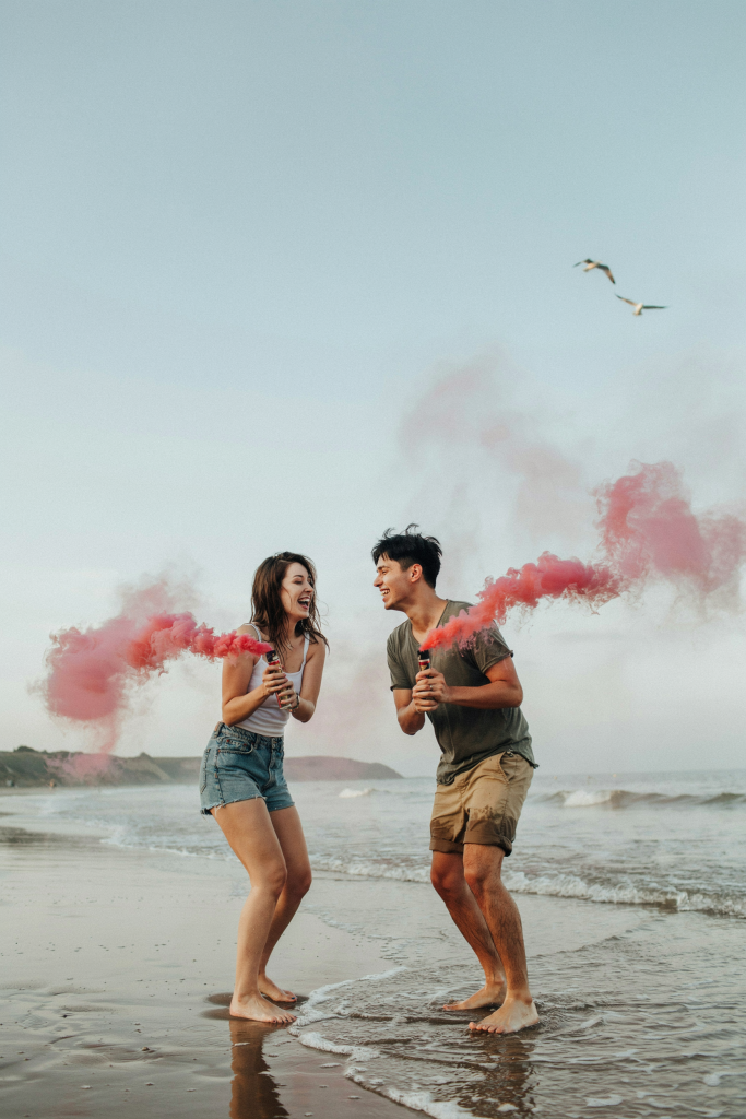 A couple standing in ankle-deep water, holding smoke bombs — one in each hand — releasing thick pink smoke into the ocean breeze. The smoke trails dramatically behind them in both directions. Faces turned toward each other, wide smiles. Shot at a slight distance so the full smoke trail is visible.