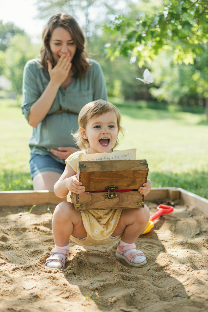 A toddler girl crouching in the sand, holding open a small wooden treasure chest. Inside the chest is a folded note that reads "You're going to be a big sister!" The little girl is caught mid-read, mouth open in delight. Mom is visible in the blurred background, hand over her mouth. Bright natural light, mid-morning
