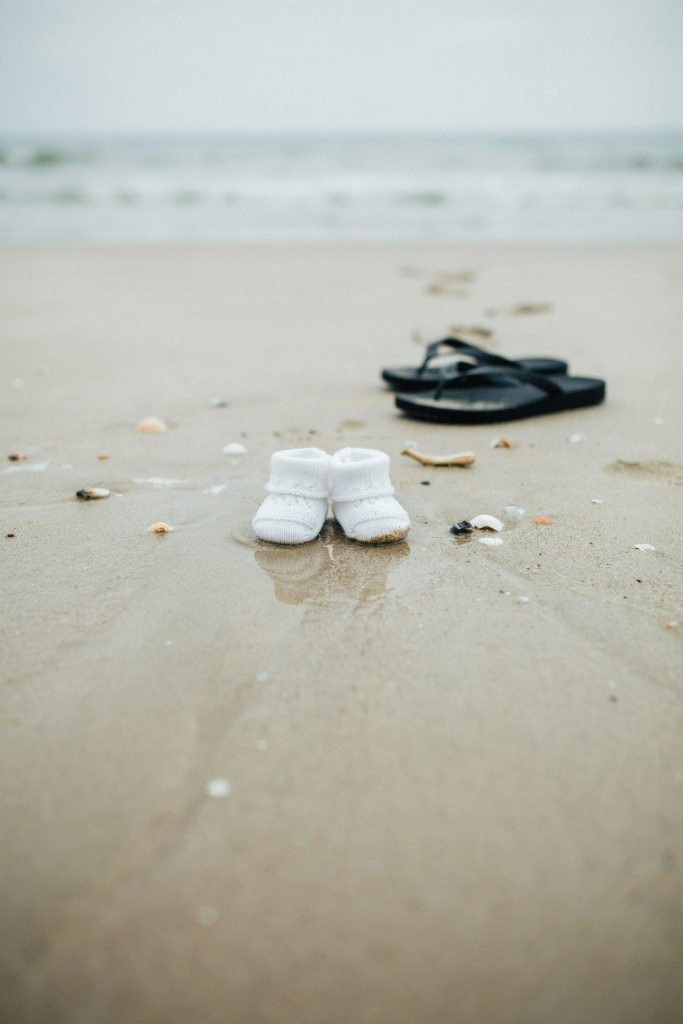 A pair of tiny white baby booties placed in the wet sand, right at the water's edge, facing the ocean. Two large adult flip-flops visible further up the beach behind them. Shot at a low angle so the booties are in sharp focus and the adults are beautifully blurred in the background.