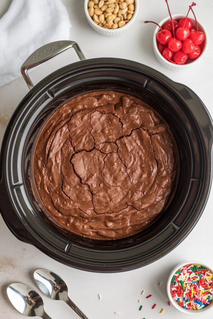 Crockpot hot fudge brownie base with glossy chocolate surface and sundae toppings arranged beside it.