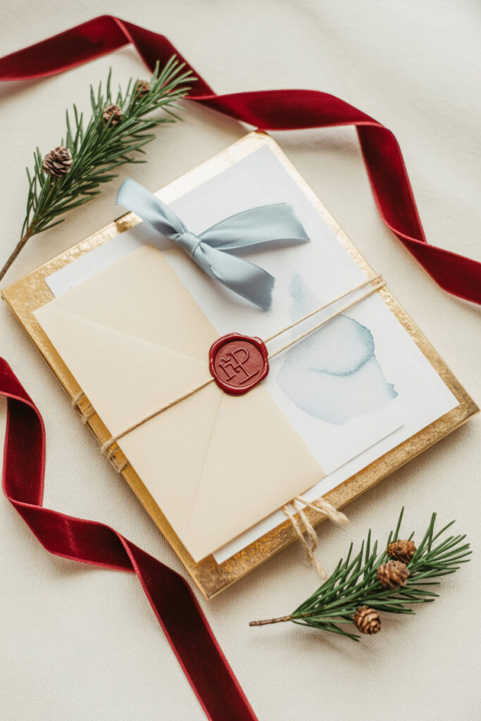 “Elegant holiday flat-lay featuring cream envelopes wrapped with twine and a red wax seal, soft blue ribbon, evergreen sprigs with tiny pinecones, and deep red velvet ribbon on a neutral background.”