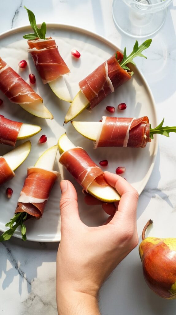 Elegant pear slices wrapped in prosciutto with fresh arugula, arranged on a white plate and garnished with pomegranate seeds for a festive holiday appetizer.