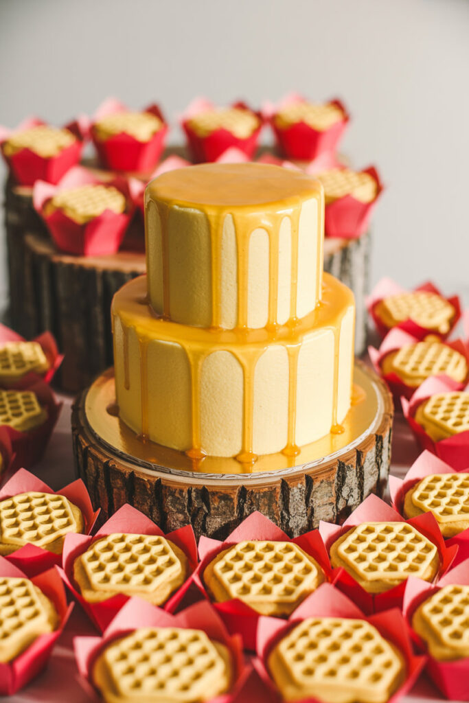 Honey-inspired dessert display featuring a yellow drip cake, honeycomb cookies, and red cupcake liners arranged on wooden risers with bright food photography.