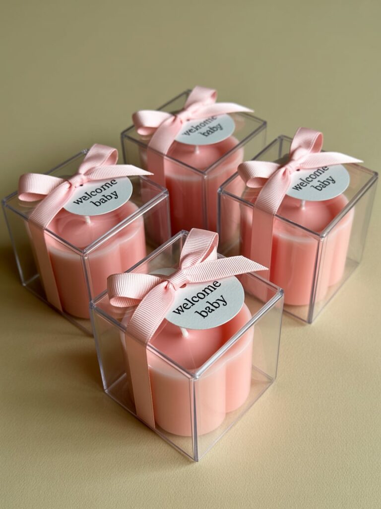 Set of mini pink candles in clear gift boxes, each tied with a blush ribbon and labeled “welcome baby” — a simple and elegant favor idea for a pink-themed baby shower.