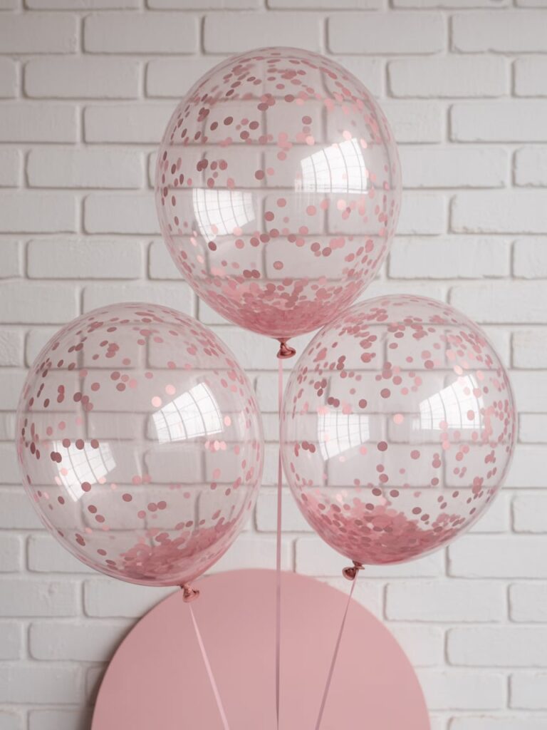 Three transparent balloons filled with pink confetti, tied with matching ribbons and set against a white brick wall — perfect for adding a festive touch to a pink-themed baby shower or party.