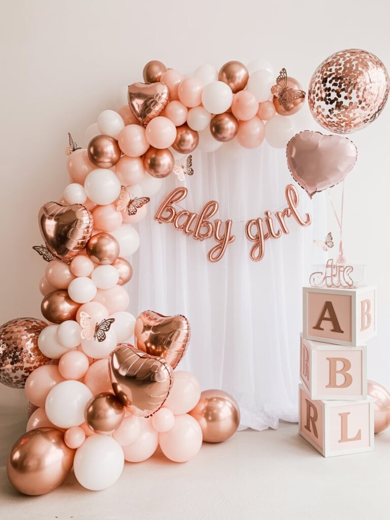 Elegant rose gold and blush pink baby shower setup featuring a balloon arch with heart-shaped metallic balloons, “baby girl” lettering, butterfly accents, and stacked alphabet blocks beside white draped fabric for a chic and modern celebration backdrop.