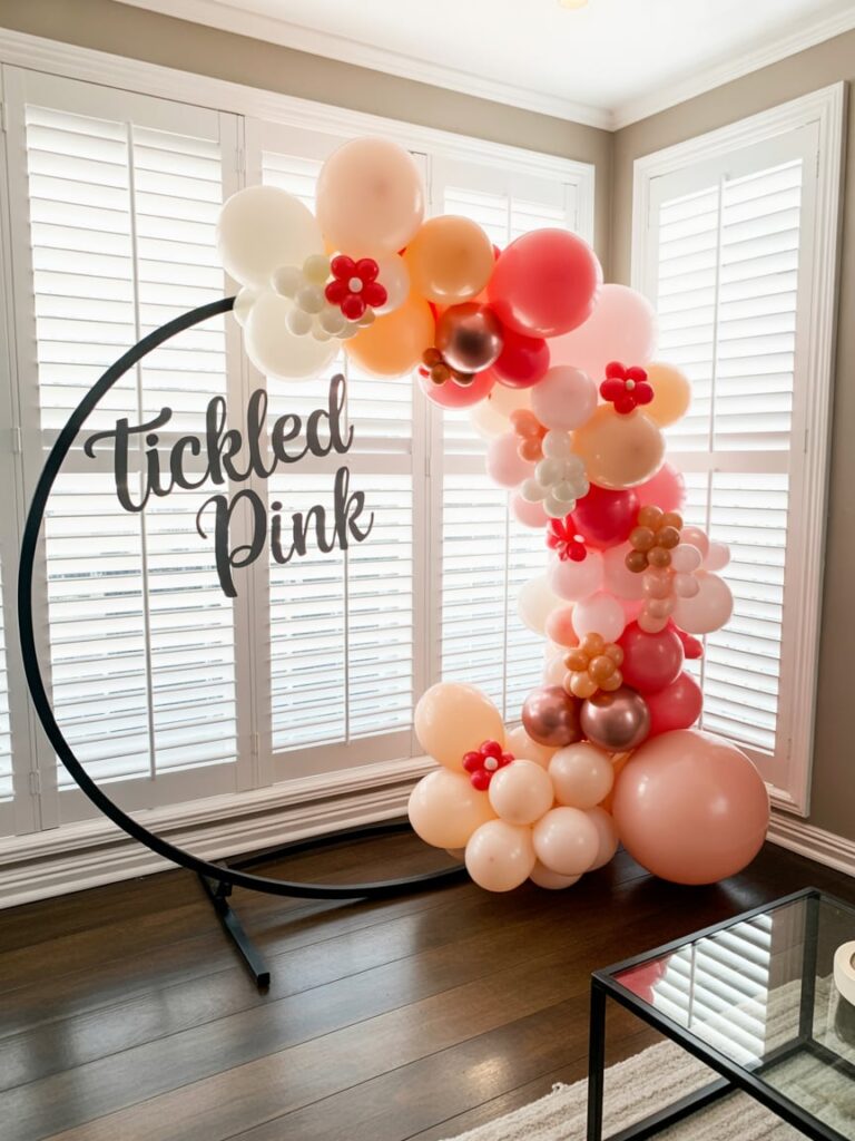 Stylish “Tickled Pink” baby shower display featuring a circular black stand adorned with pastel pink, peach, and white balloons accented with small floral balloon clusters, set against bright white window shutters for a clean and modern backdrop.