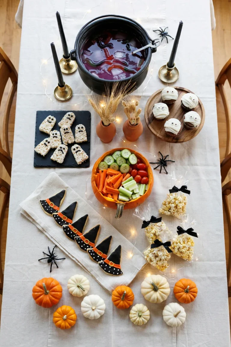 Overhead shot of a Halloween party table with mummy cupcakes, a witch's brew punch bowl, and a veggie pumpkin tray, styled with orange and black napkins and string lights