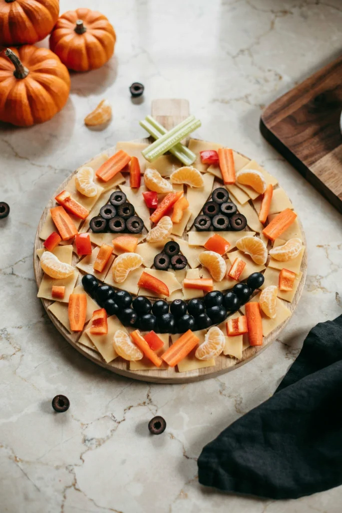 Jack-o'-lantern face snack board made of orange cheddar, clementines and carrots with black olive eyes and a dark chocolate grin, easy kid-friendly Halloween board
