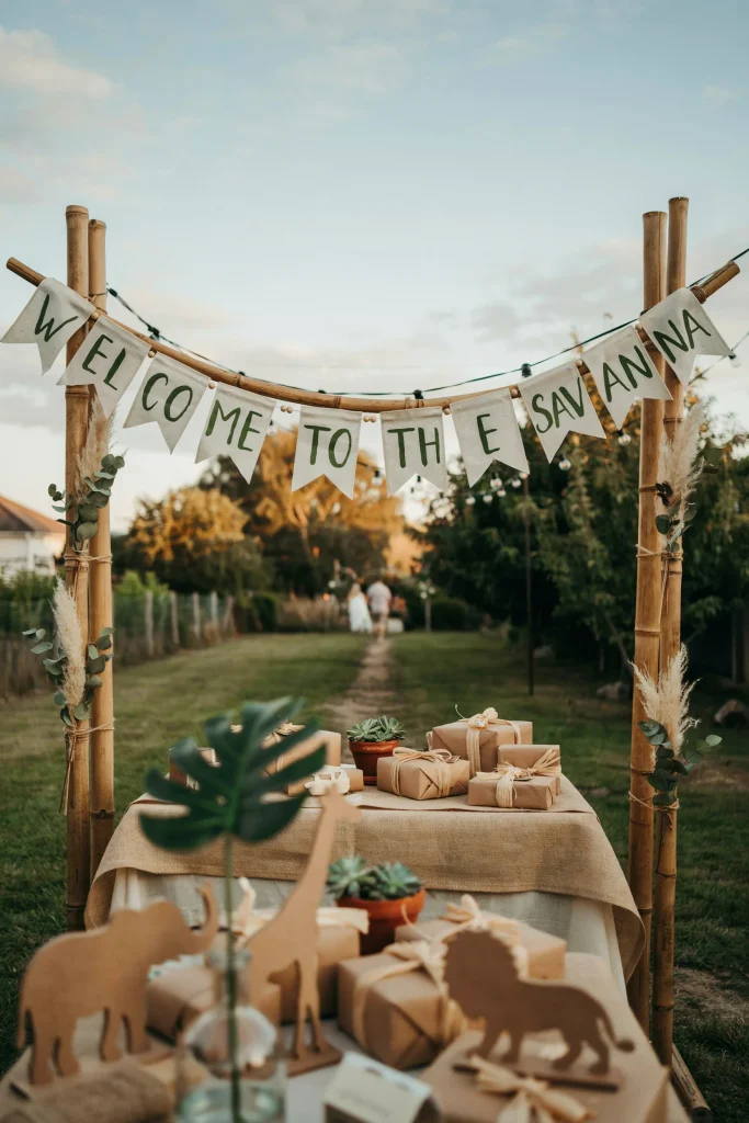 A long fabric or paper banner spelling "Safari Baby Shower" or "Welcome to the Savanna" in a hand-lettered or rustic font, strung above the entrance or behind the gift table.