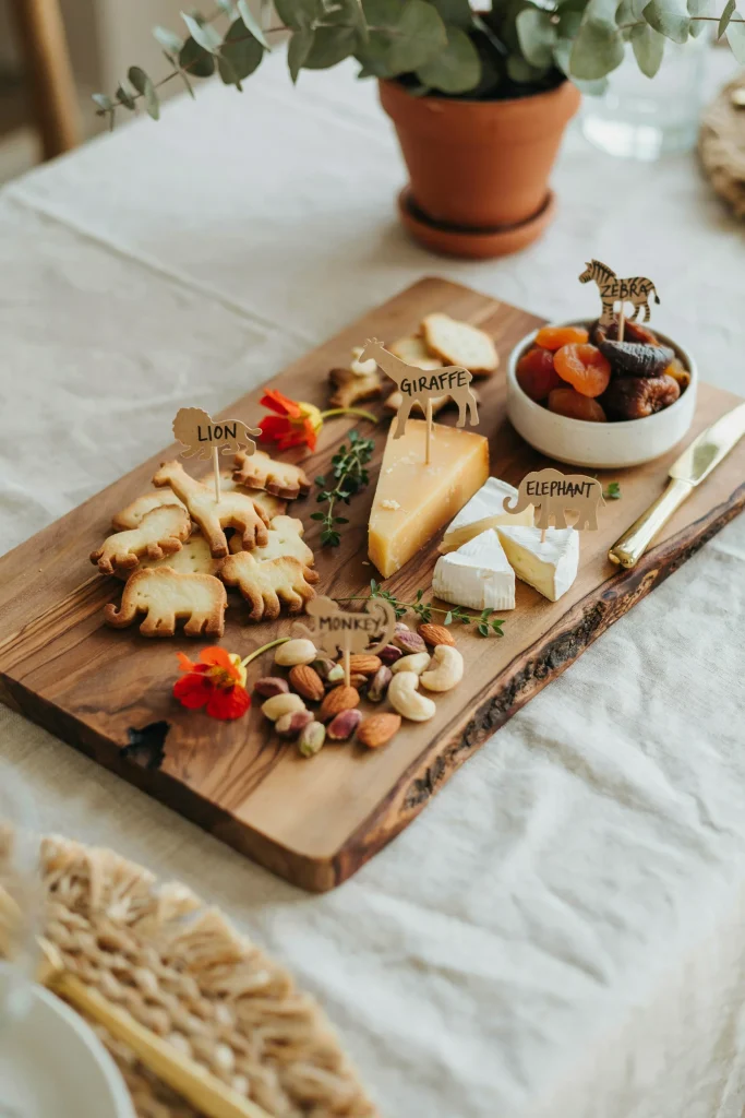 A wooden board or platter filled with animal crackers, cheese, dried fruit, nuts, and small edible flowers or herbs, arranged thoughtfully with small safari-themed labels.